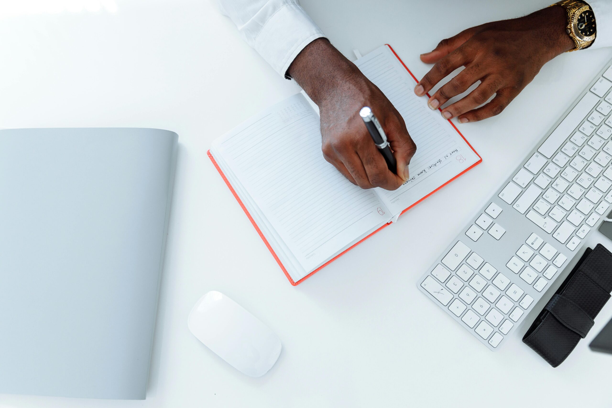 A person in a white shirt writes in an open notebook with a pen on a tidy white desk, alongside a silver keyboard, mouse, and a closed gray folder.