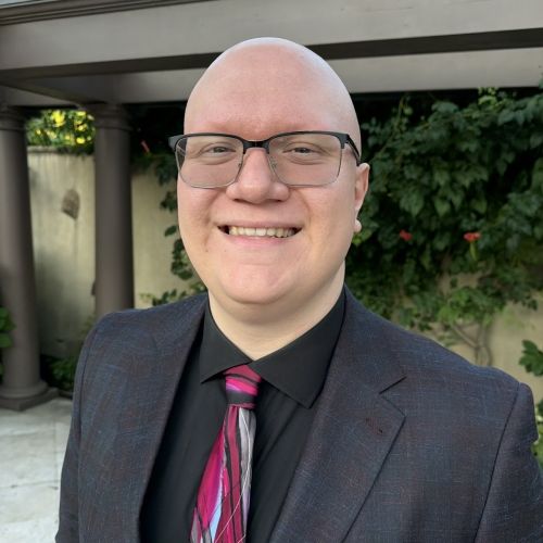 Professional headshot of Robert Petrik outdoors, wearing glasses, a dark blazer, and a pink patterned tie.