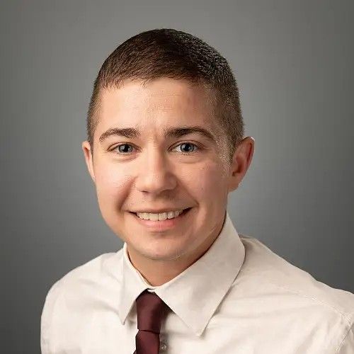 Professional headshot of Jay Harwood wearing a white dress shirt and burgundy tie, photographed against a neutral gray background.