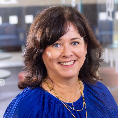 Mary Henesey wearing a blue blouse and gold necklace, photographed indoors with a softly blurred office background.